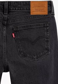 Jeans de mezclilla negra con un quinto bolsillo, parche de cuero marrón de la marca y etiqueta de logo roja en la parte trasera. Textura suave con costuras definidas.