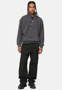 Fleece-pullover i mørkegrå med kvart-zip krage og en liten logo; parret med løse svarte bukser og svarte joggesko.