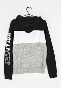 Sudadera con capucha que presenta una parte superior negra, un medio blanco y una sección inferior de color gris jaspeado. La palabra "HOLLISTER" impresa en blanco en la manga izquierda.