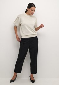 Pulover tricotat scurt cu mânecă scurtă, de culoare bej deschis, cu gât înalt și mâneci bufante, combinat cu pantaloni negri fini și sandale cu toc negru.