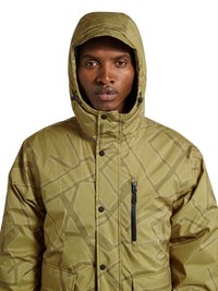 Mann trägt eine hellbraune, gemusterte Kapuzen-Winterjacke mit Reißverschlusstasche auf der Brust und Knopfleiste vorne.