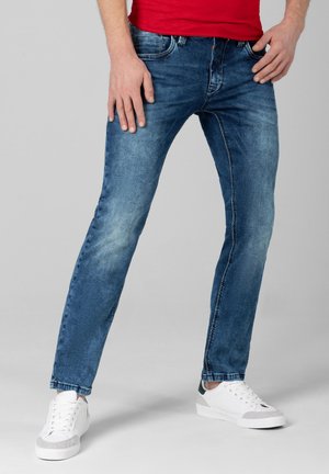 Mann trägt blaue Slim-Fit-Jeans, weiße Sneakers und ein rotes Hemd, steht mit gespreizten Beinen vor einem schlichten grauen Hintergrund.
