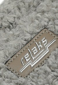 Nahaufnahme eines beigen Leder-Patches mit silbernem, geprägtem "relaks"-Logo auf einem grauen, lockeren Stoffhintergrund.