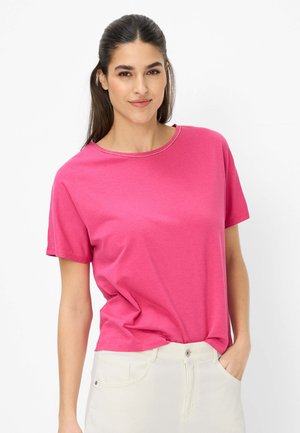 Frau mit dunklen Haaren, die ein leuchtend pinkes Kurzarmshirt und hellfarbene Hose trägt, steht mit einer Hand in der Tasche vor weißem Hintergrund.