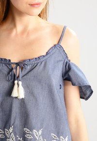 Blå chambray-top med flæseærmer, bindebånd ved halsen og hvide kvaster. Har broderede bladmønstre ved kanten. Blød tekstur.