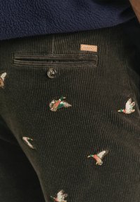 Donker corduroy broek met geborduurde vliegende eenden en een achterzak met een knoop en een klein leren GANT logo label.