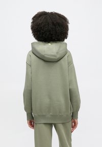 Mikina s kapucňou v svetloolivovej farbe, vyrobená z mäkkého materiálu, s voľným strihom, rebrovanými manžetami a malým neónovým akcentom na kapucni.
