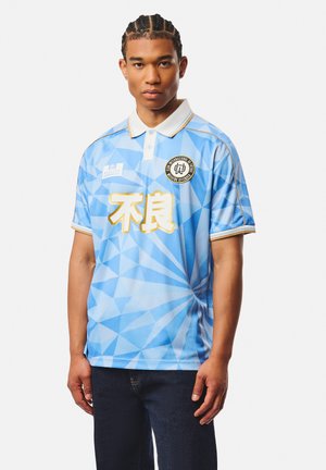 Jeune homme avec une coiffure en cornrows portant un polo bleu clair à motif géométrique avec un col blanc et des logos et textes aux accents dorés.
