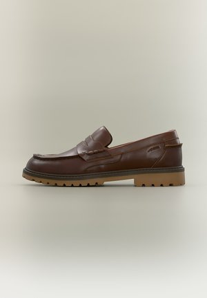 Mocasín de piel marrón con suela de goma gruesa y estriada y detalle de correa para moneda, mostrado de lado sobre un fondo liso.