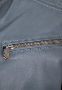 Nahaufnahme eines metallenen Reißverschlusses mit einem Zipper an weichem blauen Lederstoff, der entlang der Kanten detaillierte Stichverzierungen aufweist.