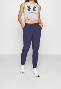 Kvinna klädd i marinblå Under Armour-joggingbyxor, vita sneakers och en knuten grå Under Armour-tanktopp, stående med händerna i fickorna.