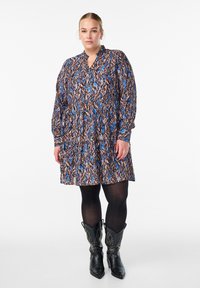 Pitkähihaisten mekko, jossa on värikäs, abstrakti kuvio sinisen ja ruskean sävyissä, kauluskäänteet ja levenevä helma. Yhdistetty mustien niittikenkien kanssa.