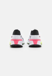 Zapatillas deportivas blancas con acentos negros, parte superior de malla texturizada, detalles en rosa y verde neón en el talón, y suela de goma negra.