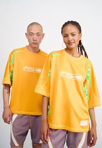 Două modele poartă tricouri atletice supradimensionate galbene cu accente verzi și un logo. Acestea sunt accesorizate cu pantaloni scurți lila cu detalii albe.