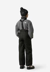 Fekete szigetelt mellesnadrág állítható pántokkal, szürke polár dzsekivel és fekete mintás kötött sapkával, sötét cipővel viselve.
