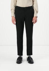 Person, der schwarze Slim-Fit-Hosen, ein beiges Hemd mit Knöpfen, weiße Socken und schwarze Lederloafers trägt, steht vor einem weißen Hintergrund.