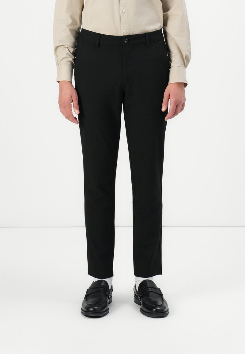 Person, der schwarze Slim-Fit-Hosen, ein beiges Hemd mit Knöpfen, weiße Socken und schwarze Lederloafers trägt, steht vor einem weißen Hintergrund.