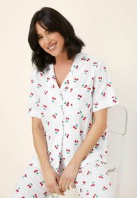 Glimlachende vrouw met schouderlang zwart haar, gekleed in witte pyjama met korte mouwen, bedrukt met rode kersen, zittend op een witte stoel.