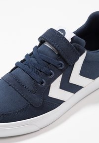 Navy canvas sneaker with white accents, featuring laces and a hook-and-loop strap. Rubber sole and textured fabric for added grip.