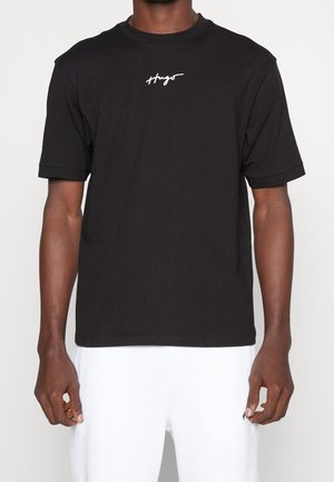 Person iført en sort T-shirt med korte ærmer med det hvide "Hugo" tekstlogo på brystet og hvide bukser, stående mod en hvid baggrund.