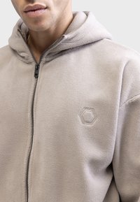 Light gray fleece zip-up hoodie with a hood, featuring a textured surface and a subtle hexagonal logo on the chest.