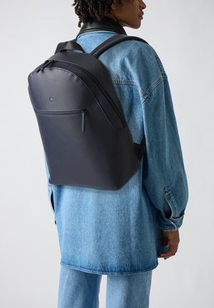 Mochila negra y minimalista con un acabado de textura suave, que cuenta con un bolsillo frontal con cremallera y correas ajustables. Llevada sobre una chaqueta de mezclilla.