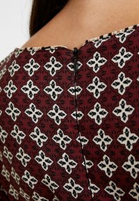 Tissu à motif rouge avec un design géométrique répété en noir et blanc, présentant une fermeture éclair noire visible au niveau du col arrière.