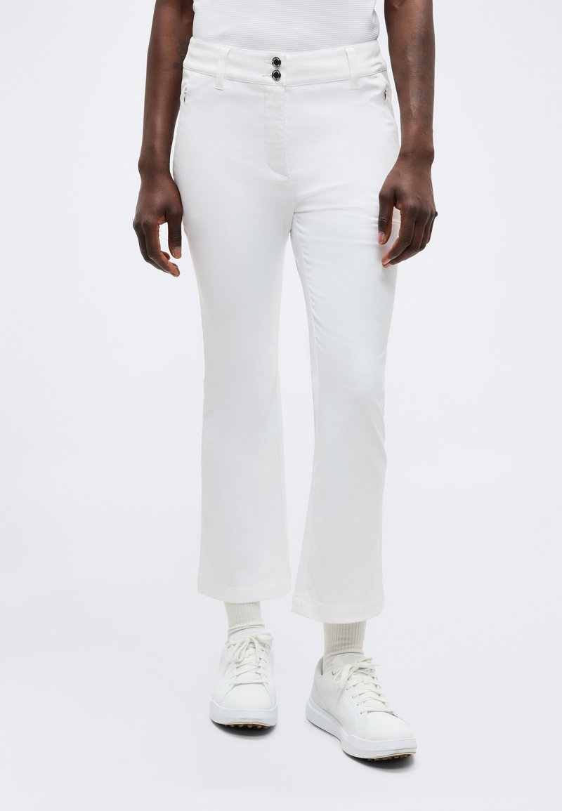 Άτομο που φοράει λευκό κοντό παντελόνι με μαύρα κουμπιά, λευκές κάλτσες και λευκά αθλητικά παπούτσια, στέκεται μπροστά σε ένα μονόχρωμο φόντο.