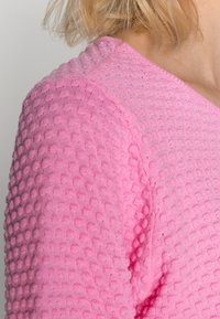 Gestrickter rosa Pullover mit strukturiertem Noppenmuster, V-Ausschnitt und weichem, leichtem Material. Der Fokus liegt auf dem Schulterbereich.