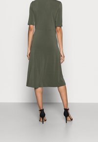 Olivgrön, knälång klänning med korta ärmar, åtsittande bodice och utsvängd kjol. Bärs med svarta högklackade sandaler. Slät textur.