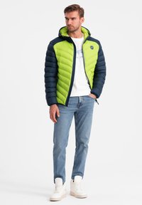 Vuorenhuppu, jossa on limevihreä etuosa ja tummansiniset hihat, vetoketjusuljenta ja tikattu pinta, yhdistettynä farkkuihin.