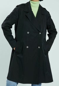 Czarny dwurzędowy płaszcz trench z dużymi błyszczącymi guzikami, kieszeniami po bokach i kołnierzem, nałożony na zielony golf. Gładka tekstura materiału.