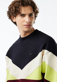 Joven con cabello rizado que lleva un sudadera de bloques de color en secciones de azul marino, crema, amarillo y marrón sobre un fondo blanco.