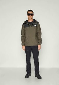 Groene en zwarte windbreaker met capuchon, ritsluiting en logo op de borst; gecombineerd met zwarte broek en robuuste zwarte schoenen. Zonnebril gedragen.