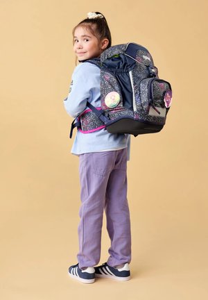 Young girl wearing a large galaxy-patterned backpack with cartoon badges, smiling and standing against a beige background.