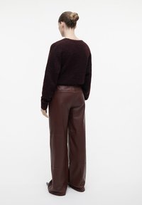 Vrouw met haar in een knot, gekleed in een donkergrote textuur trui en hoge wijdvallende bruine leren broek, staande met de rug naar de kijker gewend.