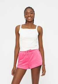 Χαμογελαστή γυναίκα με πλεξούδες, φορώντας λευκό αμάνικο τοπ και φωτεινά ροζ σορτς με κορδόνι, μπροστά σε απλό λευκό φόντο.