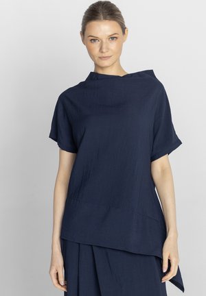 Kobieta o jasnej karnacji i włosach związanych w kok, ubrana w luźny, ciemnoniebieski top z krótkim rękawem oraz pasującą spódnicę na jasnym tle.