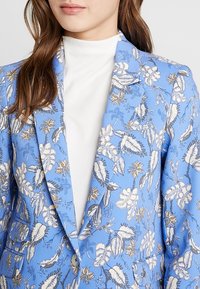 Light blue blazer with floral and leaf pattern in white and gold accents. Features structured lapels and single-button closure.