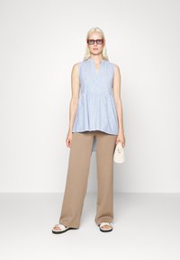 Top sem mangas azul e branco em riscas, com uma abertura de botões e design peplum, combinado com calças largas beje e sandálias brancas.