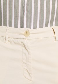 Närbild av midjan på beiga byxor med knapp och bältesöglor, tillsammans med en insydd skjorta med vertikala gröna, vita och rosa ränder.