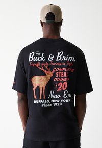 Μαύρη βαμβακερή μπλούζα με γραφικό σχέδιο που απεικονίζει έναν ελαφό, κείμενο σε κόκκινο και άσπρο. Κοντά μανίκια, χαλαρή εφαρμογή, απλή πλάτη, κανονικός λαιμός.