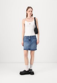 Débardeur blanc avec logo rouge, jupe en denim bleu avec ourlet effiloché, chaussures noires épaisses et chaussettes blanches. Tenue simple et décontractée.