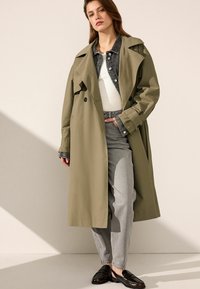 Trench coat verde oliva con bottoni doppi, indossato sopra una giacca di jeans e una maglietta bianca; abbinato a pantaloni grigi chiari e scarpe nere.