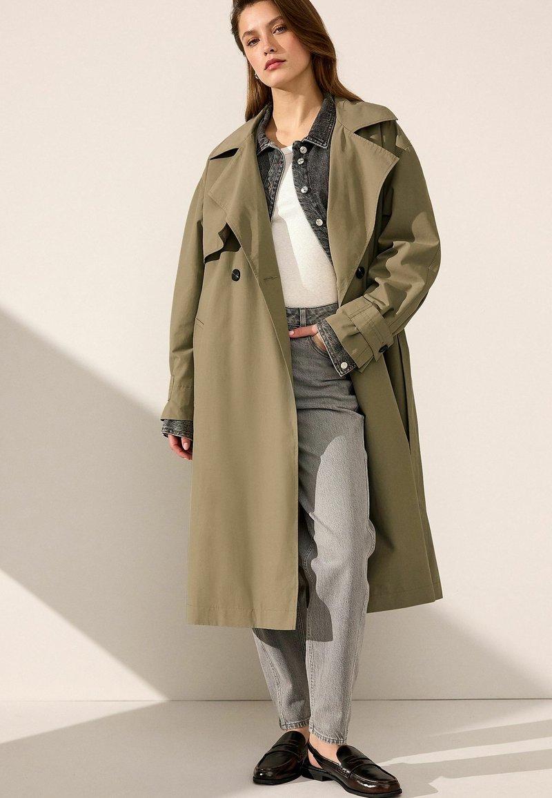 Trench coat verde oliva con bottoni doppi, indossato sopra una giacca di jeans e una maglietta bianca; abbinato a pantaloni grigi chiari e scarpe nere.