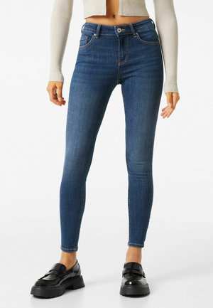 Vrouw draagt hoge taille donkerblauwe skinny jeans, cropped beige geribbelde trui en zwarte chunky loafers, staand tegen een witte achtergrond.