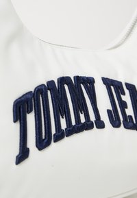 Baltas bikini viršus, kuriame yra tamsiai mėlynas siuvinėjimas "TOMMY JEANS" logotipo, lygus audinys ir apvalus iškirpimas.