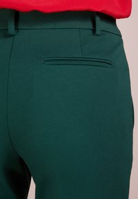 Zelené kalhoty na míru s hladkým povrchem, s kapsou vzadu a poutky na opasek, doplněné červeným topem nahoře.