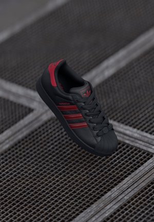 Baskets Adidas Superstar noires avec des bandes rouges et des détails, positionnées au-dessus d'un sol en grille métallique avec des lignes diagonales.