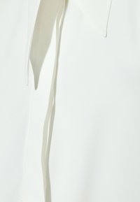 Blusa bianca realizzata in tessuto leggero e liscio, con colletto a punta e un lungo dettaglio a cravatta sul lato sinistro.